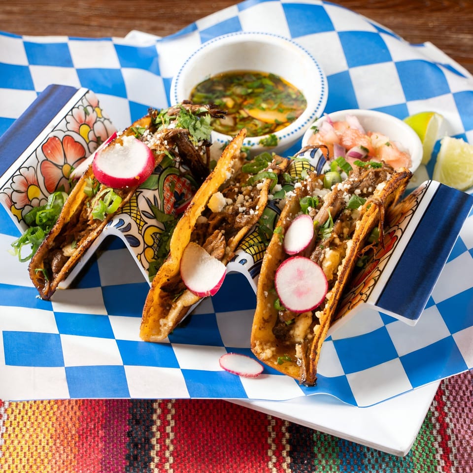 Three Tipsy Tequila and Taco Bar tacos with radishes, cilantro, and sauce on a blue checkered paper.