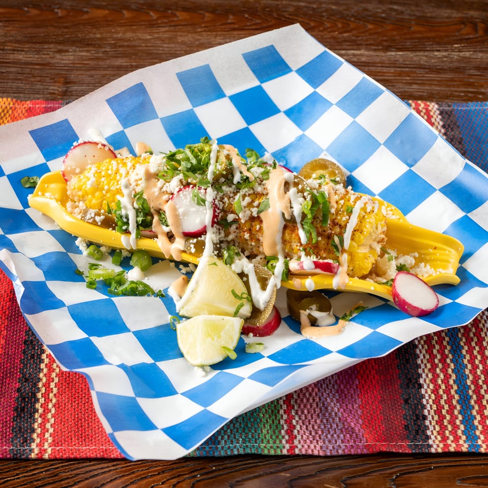 Elote with cotija cheese, crema, cilantro, radishes, and lime on a blue and white checkered paper.