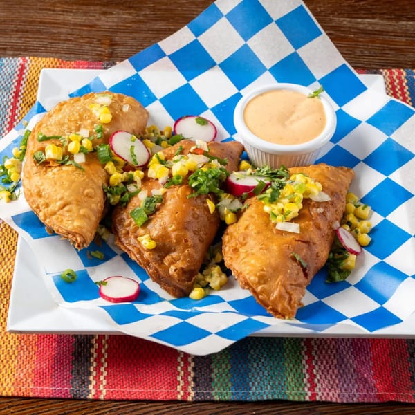 Three golden-brown empanadas topped with corn, radishes, and cilantro, served with a side of sauce.