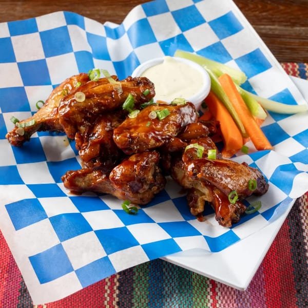Glazed chicken wings with green onions, a side of ranch, carrots, and celery on checkered paper.