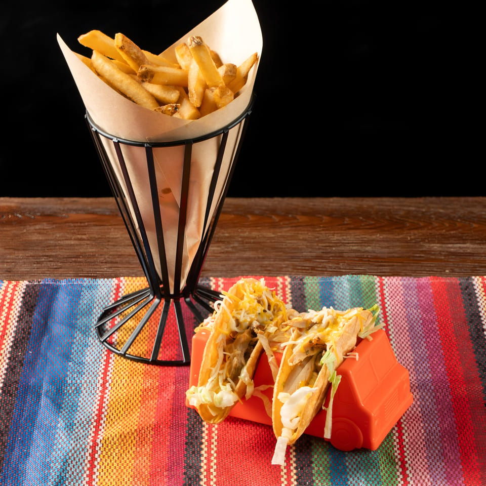 Two tacos in a red holder and a cone of french fries on a colorful striped mat.