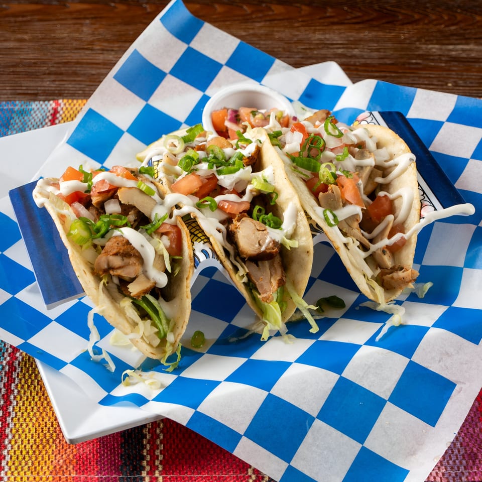 Three chicken tacos with lettuce, tomatoes, green onions, and white sauce on blue checkered paper.