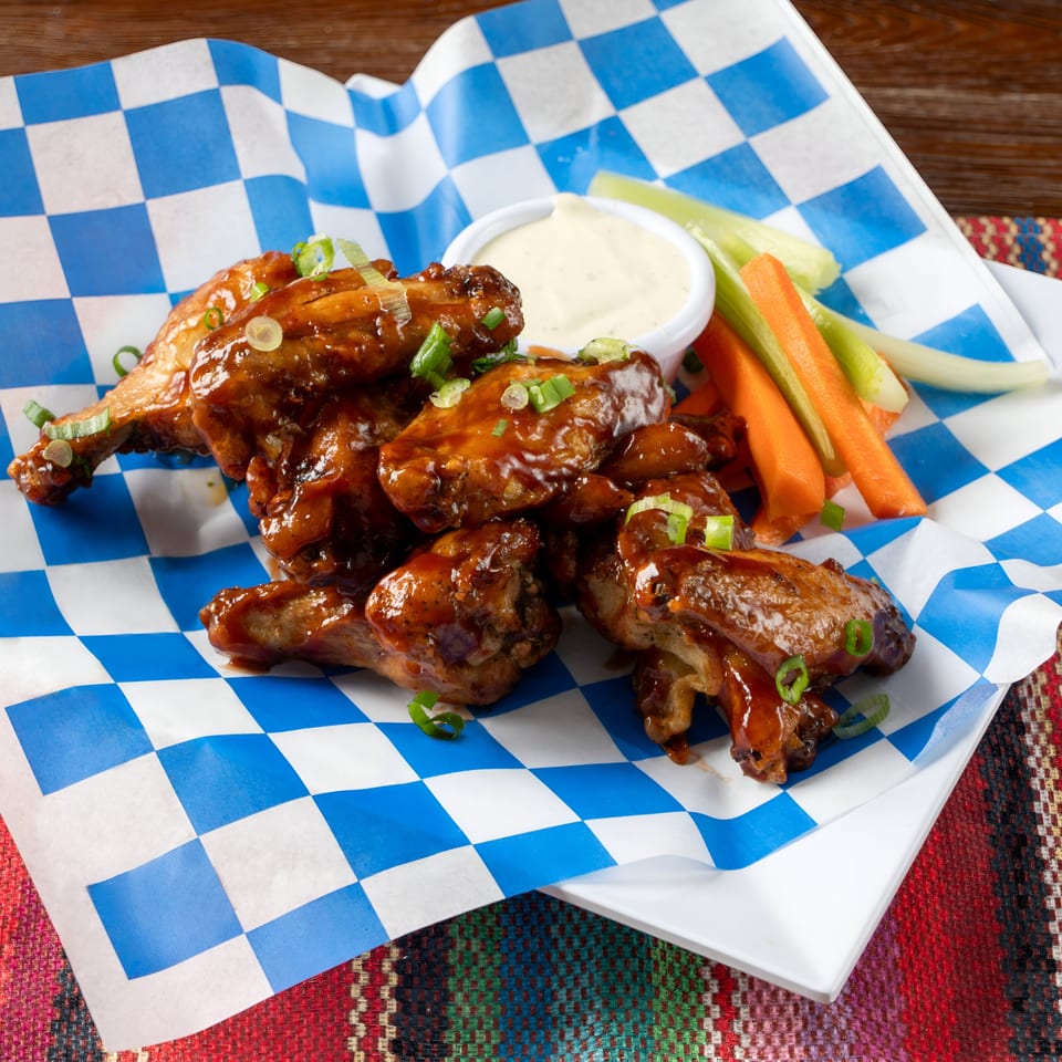 Glazed chicken wings with green onions, a side of ranch, and vegetable sticks on checkered paper.