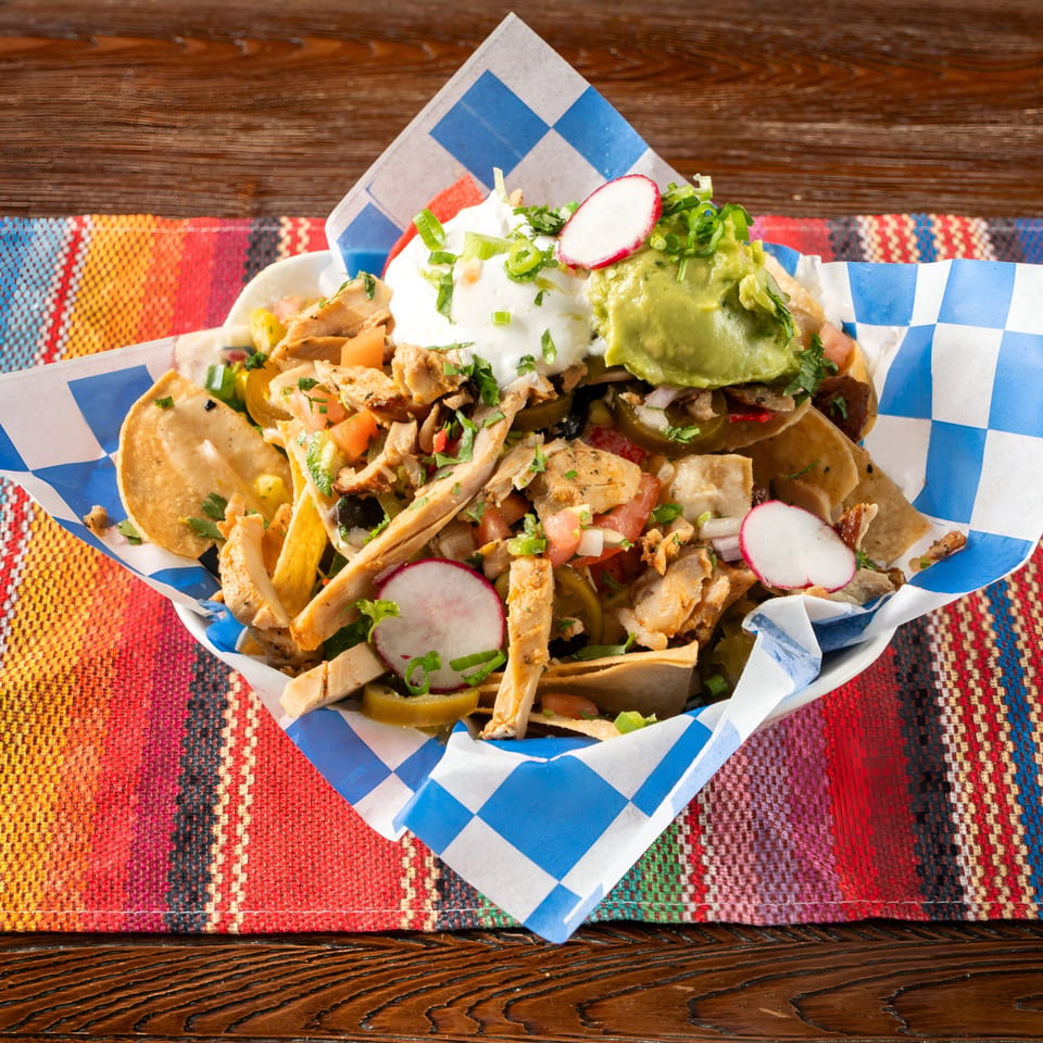 Chicken nachos with sour cream, guacamole, radishes, and jalapeños in a checkered paper-lined basket.